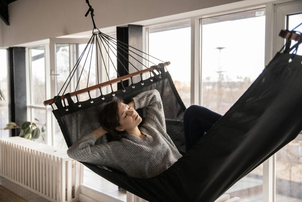 Young lady in casual wear lying in comfortable hammock leaned on hands near window and radiator in apartment in afternoon